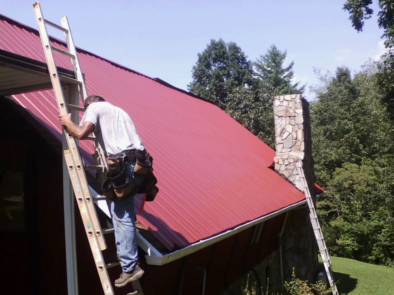 Roofer on a ladder beside a red metal roof and stone chimney — Soffit & Fascia Repair by Roofing Brothers in Mount Holly